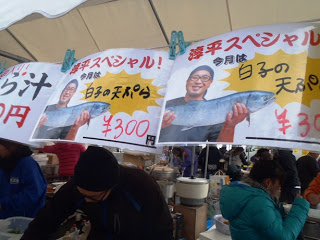 「今月は白子の天ぷら 300円」と書かれ魚を持っている男性の写真の価格ポップがテント前に並べて掲示されている出店を写した写真