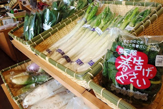 竹のふちどりがされたかごに、太ねぎや大根、袋に入った「春告げやさい」などの新鮮な野菜が並んでいる、明るい店内の野菜コーナーの写真