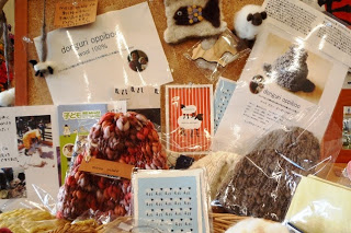 カラフルな毛糸の帽子や羊毛フェルトのマスコット、小さなカードや説明書きなどがコルクボードの前に並べて展示されている、手作りの温かみが感じられる小物コーナーの写真