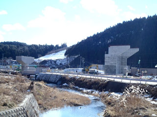 川の近くに位置する志津川地区の小森周辺で、橋脚や構築物が目立ち始め、現場には複数の建設車両や機材が並ぶ、道路工事現場の様子を写した写真