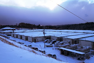 白い屋根が連なり、車や設備が整然と並んでいる雪に覆われた広大な仮設住宅地を高台から見下ろした構図の写真