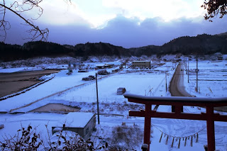 雪が積もった田畑と民家が広がる集落を、手前の赤い鳥居越しに高台から見下ろしており、静かな冬の風景が広がっている写真