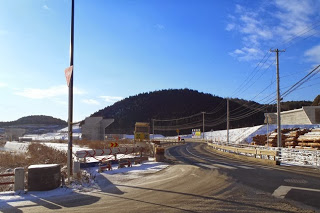 道路脇に雪が積もり、右手には木材が積まれ、左奥には盛土工事中の高架橋の橋脚が見える、青空の下の冬の道路の写真
