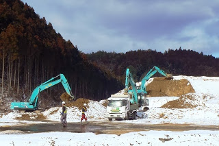 雪が積もった造成地で、重機が土をすくいダンプカーに積み込む作業を行っており、2人の作業員が見える様子の写真