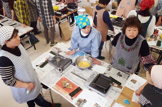 数人ずつのグループに分かれて調理台を囲み、卵焼きの準備をしている女性たちの写真