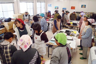 エプロンや三角巾を着けた多くの女性たちが室内に集まり、調理体験の説明を受けている写真