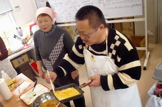 白いエプロンを着用した男性がフライパンで卵焼きを作っている様子を、女性が見守っている写真