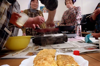 参加者たちがフライパンを使って調理する様子の前に、皿に盛られた卵焼きが写っている写真