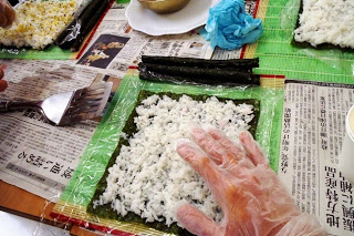ビニール手袋をした手で、海苔の上に酢飯を広げて巻き寿司を作っている途中の写真
