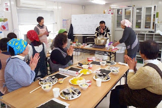 調理を終えた参加者たちがテーブルを囲んで手を合わせ、「いただきます」の挨拶をしている写真