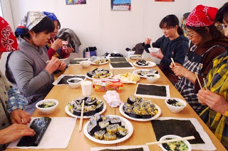 巻き寿司や味噌汁などが並ぶテーブルで、参加者たちが楽しそうに食事をしている写真