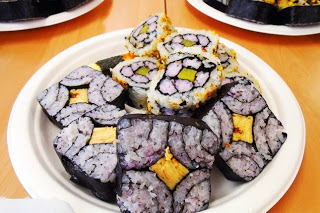 紫色の酢飯や卵焼きが入った飾りのり巻きが、白い皿に美しく盛り付けられている写真