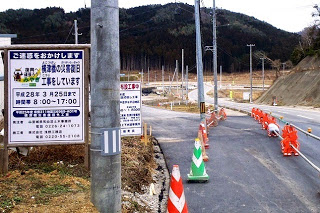 舗装工事中の道路にカラーコーンが並べられ、左側の看板には工事期間や作業時間が書かれている写真