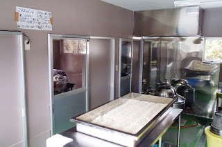 ステンレス製の設備や作業台が並ぶ調理場で、手前のテーブルにはおからの様な大量の粉状の物が平らにならされている大きなトレーが置かれており、奥には部屋につながる扉や調理器具が見える工房内の写真