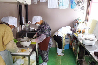 調理器具や容器、設備が棚やカウンターに並んでいるキッチン内で、白い帽子とエプロンを着た3人の女性がキッチンで作業している様子の写真