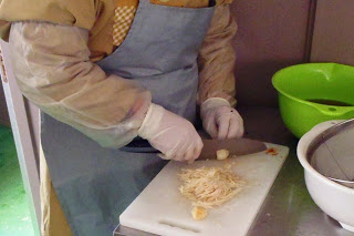 ざる付きの緑色のボウルと白いボウルが置かれたステンレス製の台の上で、手袋・エプロン・腕カバーを着用した人物が、白いまな板の上で食材を細かく切っている手元をアップで撮影した写真