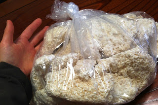 人の左手が写っている横に、白くて米の様な形をした麹が沢山詰められた、袋の口が縛られた大きなビニール袋がテーブルの上に置かれている写真