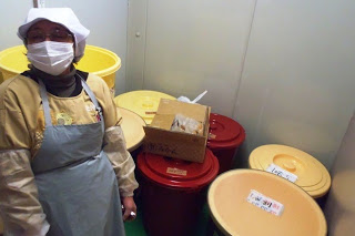 狭い空間に白い帽子と灰色のエプロンを着た女性が室内に立っており、周囲には黄色、赤色、ベージュ色の大きな容器が2つずつ置かれている写真