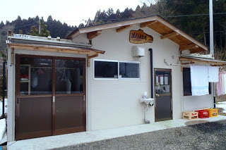 左側にはガラス戸、中央には小さな窓とドアがあり、右側には洗濯物が干されている様子が見える、白壁で屋根の下中央に「ぬくもり工房」と書かれた木製看板が設置された、木造の小さな建物外観の写真