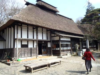 茅葺き屋根の伝統的な日本家屋の前に木製のベンチが置かれた砂利の庭があり、周囲には木々が立ち並ぶ中、赤い上着を着た女性が建物前の石畳の道を歩いている写真