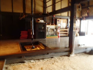 木の梁が見える趣のある建物の中で、土間から一段上がった板の間に囲炉裏がある室内の様子を写した写真