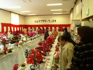 室内の広い会場の壁に紅白幕が貼られ、たくさんの色鮮やかなシルクフラワーが長机の上にずらりと並べられ、たくさんの来場者が花を眺めながら歩いているシルクフラワーフェスタの写真