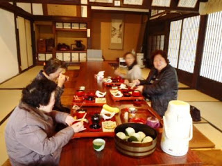 床の間に掛け軸がかけられている広々とした和室にテーブルが並んでおり、4名の女性が朱塗りのお盆にたくさんの小皿などが乗った食事を楽しんでいる様子を写した写真