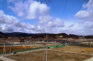 広い更地の中に道路や工事用フェンスが整備され、奥には山並みと建物が点在している風景を写した復興途中の町の写真