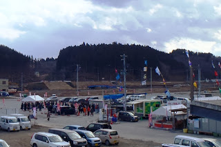 カラフルな旗がポールに掲げられた屋外の広場に集まった人々の様子が写っており、白いテントがいくつも設置された献花式の写真