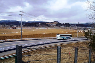 曇り空の下、田舎の風景を背景に白と青のバスが道路を走っている様子の写真