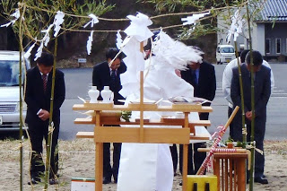 白い装束を着た神主さんが屋外の祭壇で安全祈願祭を執り行い、スーツ姿の関係者たちが深く拝礼している写真
