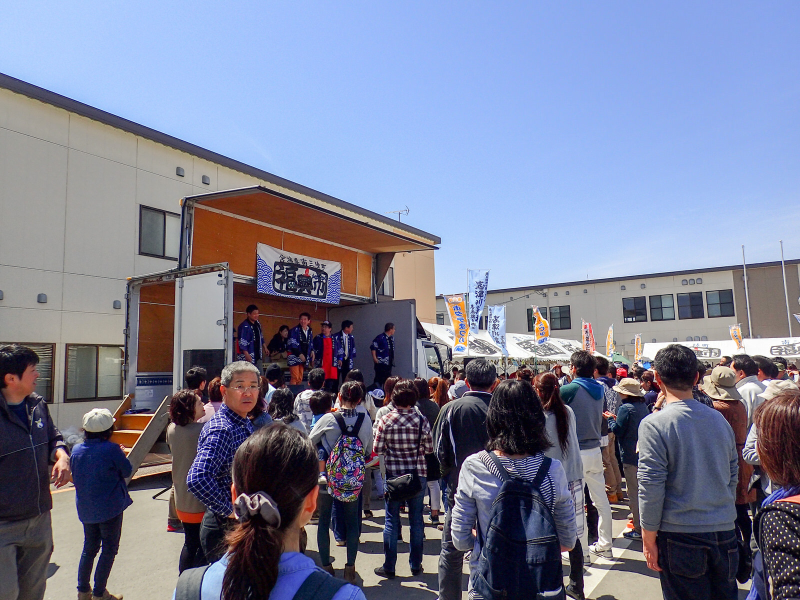出店のテントの前に立てられたたくさんののぼりが風になびく中、トラックの荷台のステージに青い法被を着た男性たちが並んで立ち、ステージの前には大勢の来場者が集まっている様子の写真