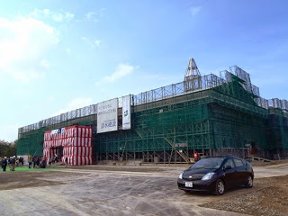 大規模な建築工事現場を捉え、全体が緑色のネットで覆われ、正面には紅白の幕がまかれ、多くの人が集まり、手前には黒い車が一台だけ止まっている写真