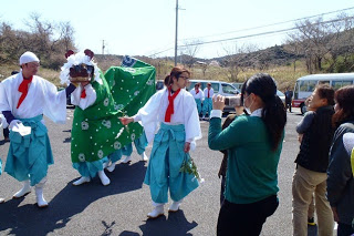 屋外で装束姿の2人に付き添われ獅子舞の被り物をしたオスッサマが歩いているところを、数人の人々が集まりビデオカメラを構えて撮影している様子の写真