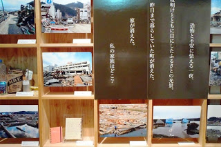 写真やメモ帳などが並んでいる木製の棚に、「恐怖と不安に怯える一夜、夜明けとともに目にしたふるさとの光景。昨日まで暮らしていた町が消えた。家が消えた。私の家族はどこ?」と書かれた黒色のパネルが設置された写真