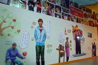 壁の上に沢山の写真が飾られ、下にはリンゴがなっている大きな木などのイラストと共に農業や飲食店で頑張っている人達が大きな写真で展示されている写真