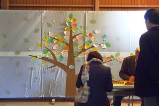 茶色の一本の大きな木が描かれた壁に、来場者による沢山のメッセージが書かれた葉っぱが貼られ、女性や男性がメッセージを記入している様子の写真