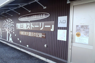 木の下に座っている子供のシルエットが大きく描かれた茶色の外壁に、今を生きる人々の言葉を伝える「南三陸ストーリー~東日本大震災の記録~」の看板が設置され、入口にポスターが貼られた建物外観の写真
