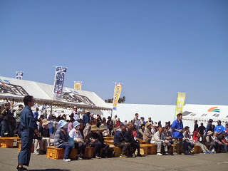 雲一つない青空の下、大きなテントの前で椅子に座った多くの人がイベントを見ている様子を写した写真