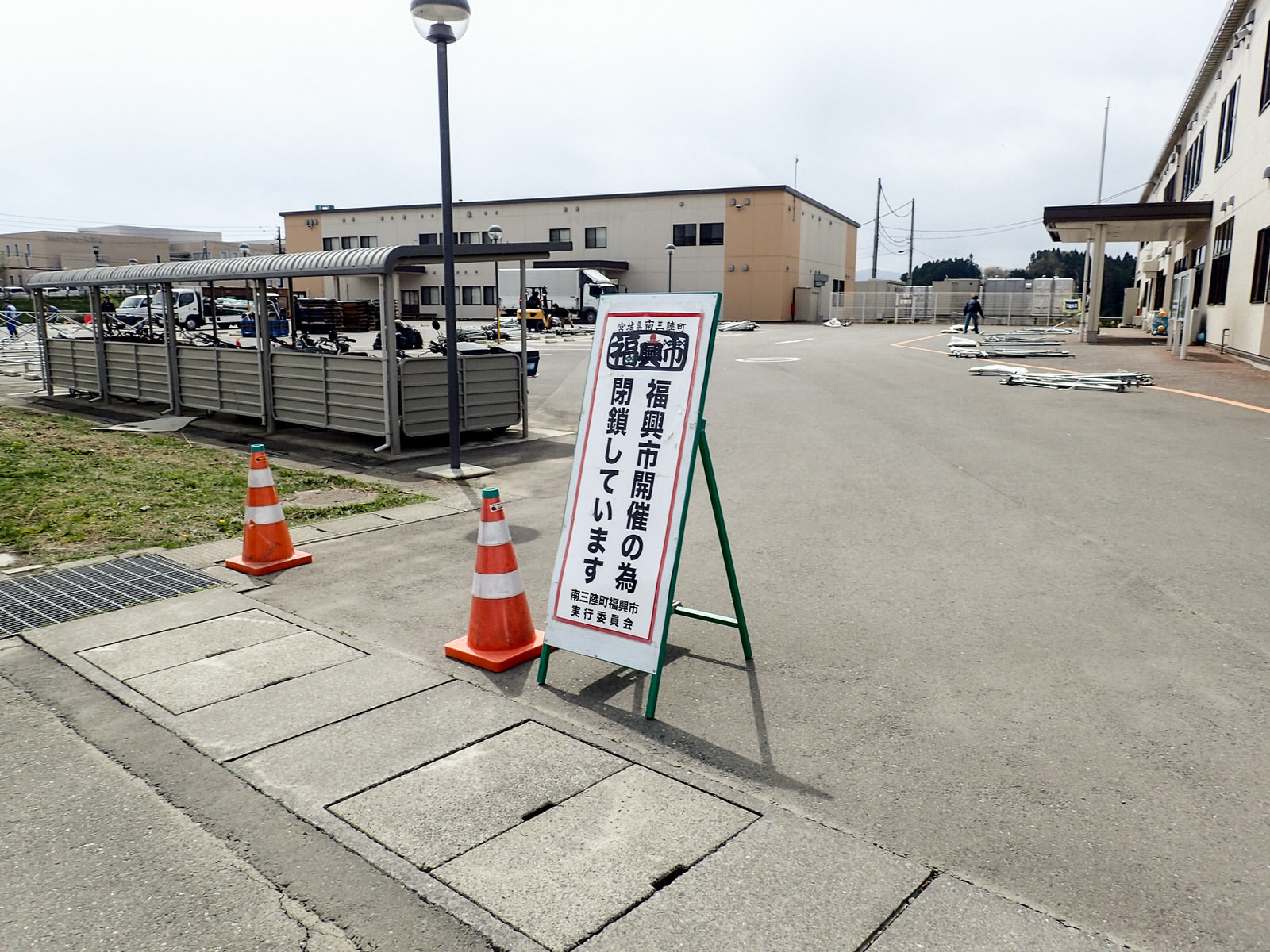 敷地の入り口に2つ並んだカラーコーンの横に「福興市開催の為閉鎖しています」と書かれた看板が立てられ、後方の駐車場では会場の準備が進む様子が見える写真