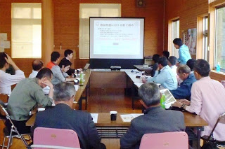 プロジェクターでスライドを映し出しながら十数人の参加者が、ロの字型に設置した長机を囲んで真剣に会議をしている写真