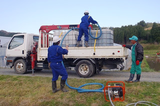 白いトラックの荷台に積まれた大型のタンクにホースをつないで液体を注入しており、作業員が3人で協力して液肥の運搬準備をしている様子を写した液肥積み込み作業の写真
