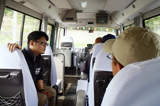 数人の参加者が座っている小型バスの中で、左側の席に座っている黒いシャツの男性が、右側を向いて話をしている写真