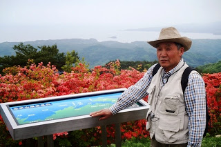 赤いツツジの花が咲く展望台で、展望パネルの横に立っている帽子をかぶった小野寺先生の写真