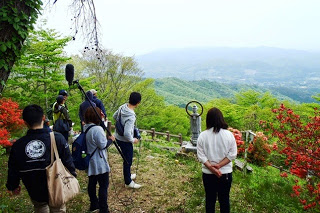 緑の山々と谷を見渡せる場所で、参加者の人たちが景色を見たり話を聞いたりしている写真