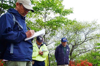 ツツジの咲く丘の上で、キャップを被った参加者たちが真剣にメモを取りながらガイドの説明を聞いている写真