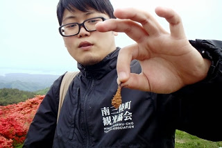 黒いジャンパーを着た男性が、小さな松ぼっくりのようなものを指でつまんでカメラに向かって見せている写真