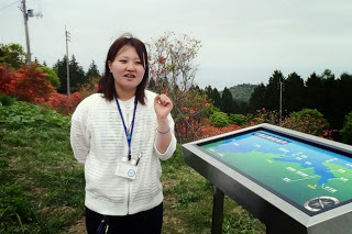 咲き誇った赤いツツジや山並みを背景に、白い服を着た女性が、展望パネルの横に立って左腕を上げて話をしている写真