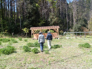 林の近くに建てられた木製の屋根付き小屋に向かって関係者2名が野原を歩いている写真