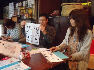 古民家の室内で人々が机を囲んで座り、紙を切って「きりこ」づくりをしており、中央の男性が持ち上げた白い紙にはお餅の模様が浮かび上がっている写真